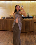 Karunia Fischer's taking a mirror selfie in a stylish, warmly lit interior with wooden floors and minimalist decor. She is wearing a long Mulberry silk aman bias skirt mud paired with a matching ecoprint scarf-style top draped elegantly around her neck and shoulders, creating a sophisticated and artistic look. Her long dark hair is worn naturally, and she poses confidently while holding her phone.