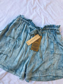 Annie Silk Shorts - Indigo TieDye by Rimmba | Sustainable Clothing feature a light blue tie-dye pattern, elastic ruffled waistband, matching fabric belt, front pockets, and attached beige tags, displayed on a white surface.