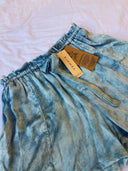 Annie Silk Shorts - Indigo TieDye by Rimmba | Sustainable Clothing, featuring an elastic waistband with drawstring, shown on a white fabric background. Two brown "RHUMAA" tags are attached to the waistband.