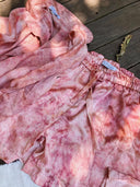 The Annie Silk Shorts - Pink Tie Dye by Rimmba | Sustainable Clothing are displayed with a matching mulberry silk button-up shirt on a wooden surface, accompanied by a dried leaf.