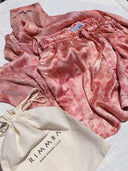 The Annie Silk Shorts - Pink Tie Dye from Rimmba | Sustainable Clothing are displayed on a white surface beside a cream drawstring bag featuring the "RIMMBA" logo and website.