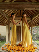 In Bali, two woman dressed with silk in a natural dye yellow; are standing on wooden pavilion with a traditional Indonesian architectural style. They are both wearing yellow Mulberry silk maxi dress with halter neck dresses and backless style. 