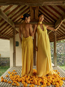 two women standing side by side in a wooden pavilion with a traditional architectural style. They are both wearing long, elegant yellow dresses that drape smoothly to the floor. One woman faces slightly away while looking over her shoulder, and the other faces forward with a calm, composed expression.