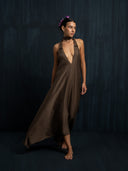 A woman stands barefoot against a dark background, wearing the flowing Aphrodite Dress - Iron Mud by Rimmba | Sustainable Clothing. Flowers and silk ribbons adorn her hair as she gazes softly to the side.