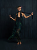 A barefoot person poses by a dark textured wall in the Aphrodite Dress - Aurora by Rimmba | Sustainable Clothing—a flowing deep green adjustable silk piece with a plunging neckline, open back, black neck ties, and pink flowers in their hair.