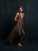 A person with short dark hair stands barefoot against a dark background, eyes closed, wearing the flowing Aphrodite Dress - Iron Mud by Rimmba | Sustainable Clothing, accented with silk ribbons and purple butterflies in their hair.