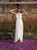 A woman in the Aphrodite Dress - White by Rimmba | Sustainable Clothing stands barefoot on purple petals, posing with her hands near her head against a rustic stone wall dotted with green plants.