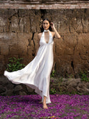 A woman in a white Aphrodite Dress by Rimmba | Sustainable Clothing walks barefoot on purple petals, framed by a textured brown stone wall and lush green plants.