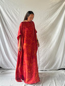 A woman stands before a white backdrop, wearing the Rimmba | Sustainable Clothing Bellas Red Silk Kaftan - Eco Print with wide sleeves and hand-dyed patterns, looking down while holding part of the dress.