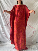 A person stands with arms slightly raised, modeling the Bellas Red Silk Kaftan - Eco Print by Rimmba | Sustainable Clothing, featuring wide sleeves and side ties. The loose-fitting design is shown against a plain draped white background; their face is partly out of frame.