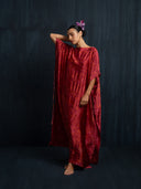 A woman stands barefoot against a dark background, wearing the Bellas Red Silk Kaftan - Eco Print by Rimmba | Sustainable Clothing. The loose, hand-dyed dress features short sleeves; she has a purple flower in her hair and looks downward.