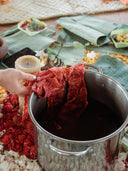 A hand holds red fabric above a metal pot of dark liquid, surrounded by petals and banana leaves, capturing the natural dyeing process at the Botanical Dye Lingerie Workshop by Rimmba | Sustainable Clothing.