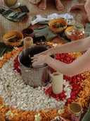 During Rimmba’s Botanical Dye Lingerie Workshop, hands rest in a metal pot among red, white, and yellow petals—possibly colored with plant dyes—candles, and jars on a woven mat. Nearby bowls and feet are also visible.