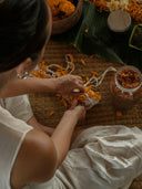 A person in a white dress arranges orange petals and white cloth on a mat, surrounded by jars and leaves—suggesting the Botanical Dye Lingerie Workshop by Rimmba | Sustainable Clothing.