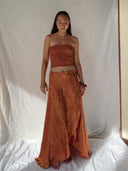 A woman with long dark hair stands barefoot on white fabric, wearing Rimmba | Sustainable Clothing's Botanical Jannah Gold Wrap Skirt and matching strapless mulberry silk top, against a white draped background. She gazes down gently.