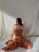 A woman with long hair sits on the floor in soft sunlight, wearing the rust-colored Botanical Silk Dayu - Elastic Bandeau Top by Rimmba | Sustainable Clothing, paired with a matching skirt and framed by a white fabric backdrop.