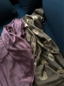 The Botanical Silk Sarong by Rimmba | Sustainable Clothing, in mauve mulberry silk and naturally dyed beige with fan patterns, is draped on a dark blue couch, creating a soft and cozy arrangement.