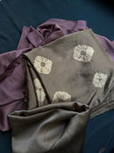 A close-up of the Botanical Silk Sarong by Rimmba | Sustainable Clothing, featuring folded, naturally dyed Mulberry Silk in muted purple and taupe tones, with one piece displaying white tie-dye style geometric patterns on a dark surface.