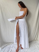 A woman stands barefoot on a white sheet, wearing the Botanical Silk Dayu - Elastic Bandeau Top by Rimmba | Sustainable Clothing and a long white skirt with a high slit, looking down and holding white fabric against a neutral draped background.