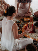During the Botanical Dye Lingerie Workshop by Rimmba | Sustainable Clothing, a woman in white sits cross-legged on a mat, gesturing as she leads a creative session surrounded by others and natural floral arrangements.