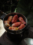 A metal steamer pot with food wrapped in reddish leaves sits on a stove. Nearby, large green leaves and blooms echo the delicate artistry of the Rimmba | Sustainable Clothing Botanical Dye Lingerie Workshop.