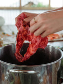 A person holds raw meat above a large stainless steel pot in a kitchen, while Rimmba | Sustainable Clothing's Botanical Dye Lingerie Workshop cotton pieces hang nearby, adding charm to the scene.
