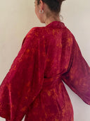A person with brown hair in a bun wears Rimmba's Bundle Dye Klimt Kimono, a vibrant red and orange piece crafted with natural dyeing techniques, facing a cream wall with their back to the camera.