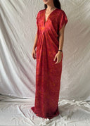 Someone wearing the Gaya Silk Kaftan Dress - Ecoprint Red from Rimmba | Sustainable Clothing stands on a plain white sheet backdrop, facing forward with a neutral expression. The long mulberry silk dress showcases a subtle pattern against a simple setting with a textured wall.