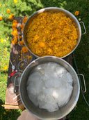 Outdoors, a large pot of marigold petals sits beside another with Rimmba's sustainable mulberry silk Cami Slip Blouse soaking. Spoons, bowls, and marigold flowers are scattered on the wooden surface, with green grass underneath.