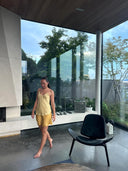 A woman in a yellow Cami Slip Blouse by Rimmba | Sustainable Clothing walks barefoot in a modern room with large glass windows, concrete floors, a black chair, and greenery outside.