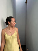 A woman in a yellow Cami Slip Blouse by Rimmba | Sustainable Clothing stands by a white wall in a modern hallway, her hair in a bun, wearing hoop earrings and a necklace. Sunlight streams from above as she looks to her left.