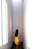 A woman in a marigold Cami Slip Blouse by Rimmba | Sustainable Clothing and a flowing skirt descends a narrow, white-walled staircase, sunlight illuminating the botanical-dyed mulberry silk. Her back faces the camera.