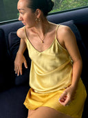 A woman in the Rimmba | Sustainable Clothing Cami Slip Blouse, crafted from mulberry silk with botanical dyes, sits on a dark sofa in sunlight. Her hair is tied back and she accessorizes with gold hoops, a necklace, and a ring.