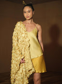 A woman stands indoors in a Rimmba | Sustainable Clothing Cami Slip Blouse and skirt, draped in a textured yellow shawl. Her hair is styled in a bun with an accessory, her look glowing with the charm of botanical dyes.