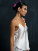 Woman wearing a white silk camisole and white silk long pants against a dark background.