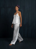 Barefoot woman wearing a white camisole silk blouse and white silk pants against a dark background.