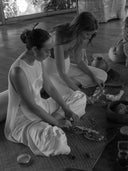 Two women sit cross-legged indoors, arranging natural materials on a mat. Surrounded by jars and bowls, they wear Rimmba | Sustainable Clothing's Cempaka blouse, enhanced by delicate Silk Satin Details for a serene atmosphere.