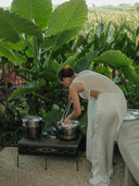 Wearing the Cempaka blouse by Rimmba | Sustainable Clothing, a woman stands by an outdoor gas stove, cooking with pots and chopsticks amid lush green tropical plants and large leaves.