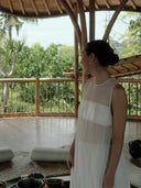 A woman wears the Cempaka blouse by Rimmba | Sustainable Clothing, standing indoors on a wooden floor with bamboo railings and a thatched roof, looking out at lush trees; yoga mats and bowls rest nearby.