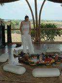 A woman in a white Cempaka blouse by Rimmba | Sustainable Clothing stands on a woven mat in an open-air bamboo pavilion, surrounded by cushions and plates of colorful food, with greenery and fields stretching beyond.