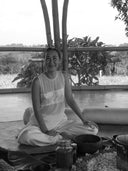 Wearing the Cempaka blouse by Rimmba | Sustainable Clothing, a woman sits cross-legged indoors, smiling amid jars and bowls. Large windows frame views of trees and fields behind her.