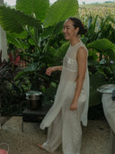 A woman in Rimmba's Cempaka blouse, a sheer white silk-chiffon top with mother-of-pearl buttons, smiles barefoot on a patio surrounded by tropical plants as a pot simmers on a small stove nearby.