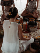 Three women sit in a serene circle on mats with flower petals and jars. One wears the elegant Cempaka blouse by Rimmba | Sustainable Clothing, featuring Silk Satin Details and flowers in her hair, adding grace to the ceremonial scene.