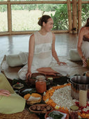 A woman in a sheer white Cempaka blouse by Rimmba | Sustainable Clothing sits cross-legged on a woven mat indoors, surrounded by jars, bowls of food, flower petals, and greenery at a peaceful group gathering.