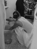 a woman crouching down indoors, washing or handling items in a large plastic container filled with water. She is wearing a white Cempaka blouse, sleeveless dress with an open back, and her hair is neatly tied up in a bun with decorative hair accessories.