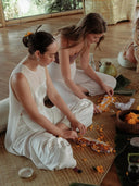 two women sitting cross-legged on a woven mat indoors, carefully preparing flower offerings. They are both dressed in flowing white outfits, creating a calm and harmonious atmosphere.