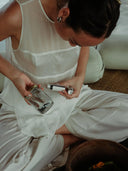 a woman sitting cross-legged indoors, wearing a soft, flowing white dress. She is focused on a small, careful task, holding a glass jar in one hand and a small bottle in the other, as if measuring or preparing a liquid.