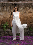 A barefoot woman standing is facing to the camera wearing Silkchiffon Cempaka Blouse white and Nindy silk pants white against a traditional clay wall.