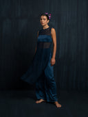 Barefoot woman wearing an indigo silk chiffon blouse and matching colour silk long pants against a dark background.