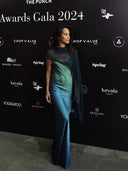 A woman in a blue-green ombre gown with a sheer dark shawl poses on a gray carpet in front of a black backdrop with "Awards Gala 2024" and various sponsor logos.